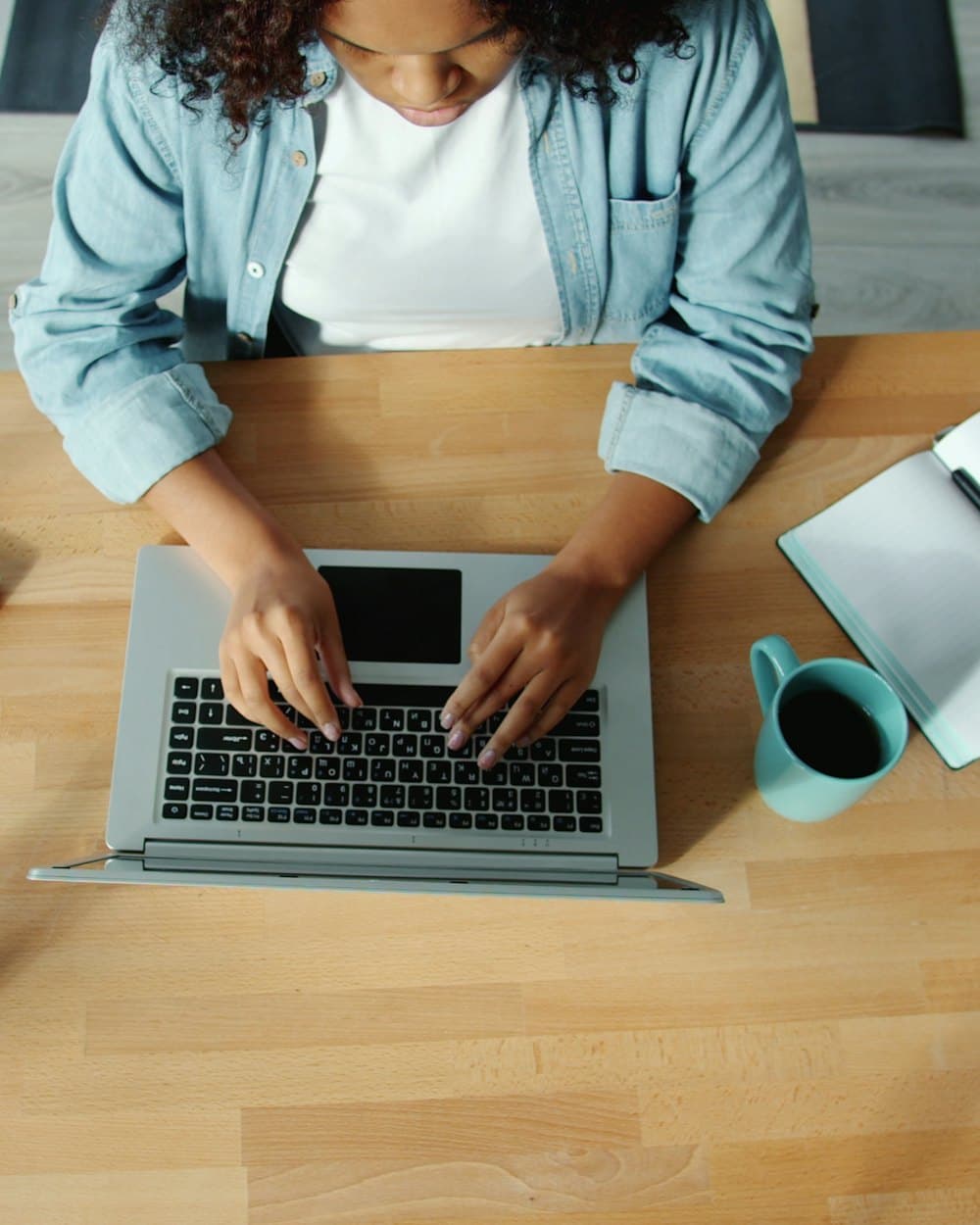 A student typing on a laptop at a wooden desk, a notebook open beside it.